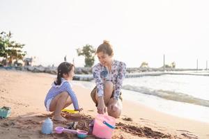 asiatico carino poco ragazza e sua madre giocando o fabbricazione sabbia castello o scavando con sabbia su tropicale spiaggia. bambini con bellissimo mare, sabbia blu cielo. contento bambini su vacanze mare su il spiaggia. foto