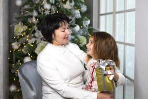 nonna con un' poco ragazza su il sfondo di Natale decorazioni e un' grande finestra. famiglia vacanza, emozioni, regalo scatola. nipotina su della nonna giro. nuovo anno foto