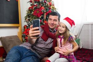 papà e figlia vicino il Natale albero con un' smartphone prendere un' autoscatto, comunicare attraverso video connessione. Natale saluti, un' regalo scatola, un' ragazza nel un' Santa cappello agitando ciao. nuovo anno, vacanza arredamento foto