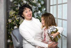 nonna con un' poco ragazza su il sfondo di Natale decorazioni e un' grande finestra. famiglia vacanza, emozioni, regalo scatola. nipotina su della nonna giro. nuovo anno foto