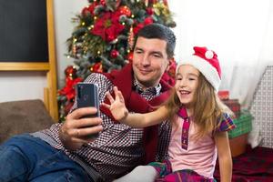 papà e figlia vicino il Natale albero con un' smartphone prendere un' autoscatto, comunicare attraverso video connessione. Natale saluti, un' regalo scatola, un' ragazza nel un' Santa cappello agitando ciao. nuovo anno, vacanza arredamento foto