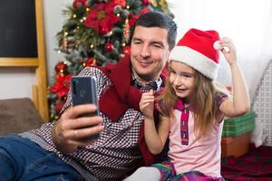 papà e figlia vicino il Natale albero con un' smartphone prendere un' autoscatto, comunicare attraverso video connessione. Natale saluti, un' regalo scatola, un' ragazza nel un' Santa cappello agitando ciao. nuovo anno, vacanza arredamento foto