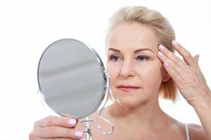 mezzo anziano donna modello toccante viso pelle guardare nel specchio. maturo più vecchio signora godendo salutare pelle cura, invecchiamento bellezza, cura della pelle trattamento cosmetico prodotti concetto. foto