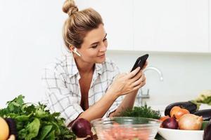 donna con senza fili auricolari è utilizzando smartphone durante cucinando nel moderno bianca cucina foto