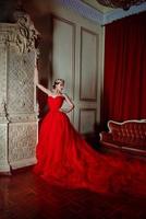 bellissimo donna nel lungo rosso vestito e nel reale corona vicino camino nel lusso interno nel foto studio