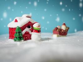 contento Santa Claus con i regali scatola su il neve slitta andando per neve Casa. vicino neve Casa avere pupazzo di neve e Natale albero. Santa Claus e neve Casa su il neve il sfondo è polvere blu. foto