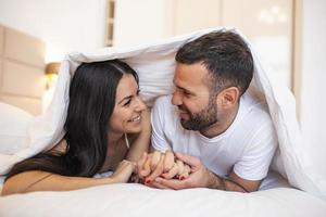 sorridente coppia andando per bacio sotto addormentato lenzuola nel il mattina. nel amore giovane contento coppia dire bugie nel letto nel il mattina foto