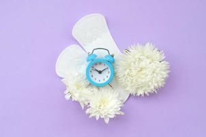 blu allarme orologio e bianca fiori bugie su mestruale pastiglie su pastello lilla sfondo foto