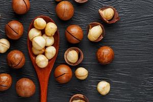 sgusciate macadamia Noce e pelato macadamia Noce su un' nero strutturale sfondo nel un' di legno cucchiaio foto