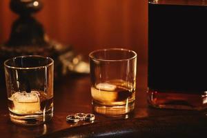 Due bicchieri di Cognac con Ghiaccio, bottiglia con modello e oro nozze anelli su un' di legno tavolo. nozze giorno foto