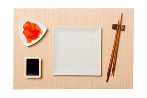 vuoto bianca piazza piatto con bastoncini per Sushi e soia salsa, Zenzero su Marrone Sushi stuoia sfondo. superiore Visualizza con copia spazio per voi design foto