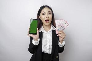sorpreso asiatico donna d'affari indossare nero completo da uomo mostrando sua smartphone e i soldi nel indonesiano rupia, isolato di bianca sfondo foto