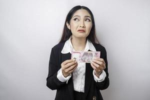 un' riflessivo giovane donna è indossare nero completo da uomo e Tenere denaro contante i soldi nel indonesiano rupia isolato di bianca sfondo foto