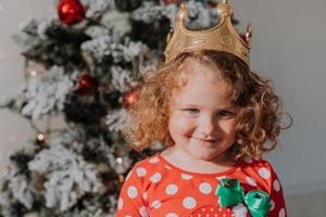 poco dai capelli ricci ragazza nel un' carnevale vestito nascosto sua viso dietro a brillante Natale albero giocattoli stelle. bambino nel un' rosso vestito con un' Santa Stampa su il sfondo di un' Natale albero. alto qualità foto