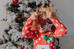 poco dai capelli ricci ragazza nel un' carnevale vestito nascosto sua viso dietro a brillante Natale albero giocattoli stelle. bambino nel un' rosso vestito con un' Santa Stampa su il sfondo di un' Natale albero. alto qualità foto