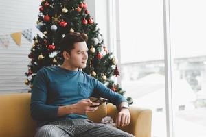 un' singolo uomo sembra solitario utilizzando il suo Telefono nel il suo vivente camera decorato con Natale alberi foto