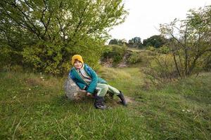 carino ragazzo sedersi su pietra a autunno collina indossare un' verde giacca, giallo cappello, con zaino e gomma da cancellare stivali. foto