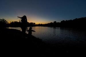 sagome di madre e figlia Tenere mano contro lago nel Alba alba. foto