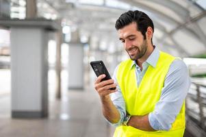 vicino su caucasico uomo uso un' smartphone per comunicare con il suo collaboratore e per contatto con il suo amico mentre lui indossare giallo veste e Lavorando a il suo luogo. foto