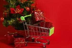 un' shopping carrello pieno di i regali contro rosso sfondo vicino piccolo Natale albero. Natale o nuovo anno shopping foto