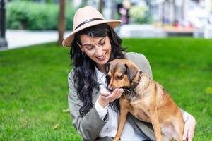 contento fricchettone donna giocando con sua cane e potabile caffè. elegante ragazza con divertente cane riposo, abbracciare e avendo divertimento nel sole, carino momenti. spazio per testo foto