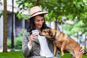 contento fricchettone donna giocando con sua cane e potabile caffè. elegante ragazza con divertente cane riposo, abbracciare e avendo divertimento nel sole, carino momenti. spazio per testo foto