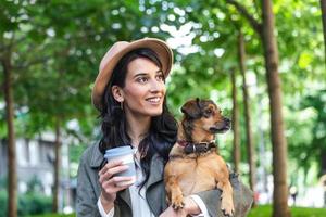 contento fricchettone donna giocando con sua cane e potabile caffè. elegante ragazza con divertente cane riposo, abbracciare e avendo divertimento nel sole, carino momenti. spazio per testo foto