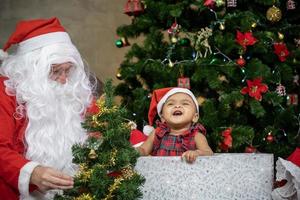 africano americano bambino è avendo divertimento ridendo e giocando con mentre Santa Claus è preparazione regalo e seduta dietro a di il Natale albero per stagione celebrazione concetto foto
