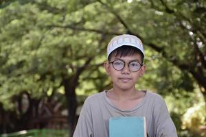 giovane asiatico musulmano ragazzo indossa occhiali, seduta nel scuola parco e lettura il suo libro nel il suo gratuito volte prima andando indietro casa, morbido e selettivo messa a fuoco. foto