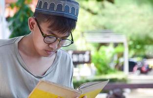 giovane asiatico musulmano ragazzo indossa occhiali, seduta nel scuola parco e lettura il suo libro nel il suo gratuito volte prima andando indietro casa, morbido e selettivo messa a fuoco. foto