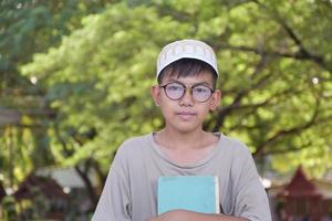 giovane asiatico musulmano ragazzo indossa occhiali, seduta nel scuola parco e lettura il suo libro nel il suo gratuito volte prima andando indietro casa, morbido e selettivo messa a fuoco. foto