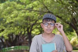 giovane asiatico musulmano ragazzo indossa occhiali, seduta nel scuola parco e lettura il suo libro nel il suo gratuito volte prima andando indietro casa, morbido e selettivo messa a fuoco. foto