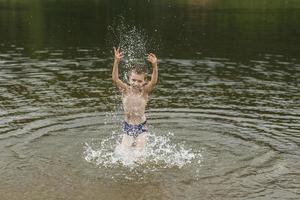 un' ragazzo avere divertimento giocando con un' spray di acqua nel il fiume a estate. copia spazio foto
