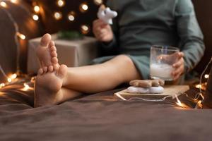 vicino su bambino spoglio piedi, bicchiere di latte cannella zabaione, natale dolci Zenzero biscotti, artigianato carta scatola regalo. ragazzo Natale mattina prima colazione. zabaione foto