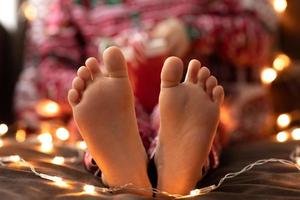 vicino su bambino spoglio piedi ragazzo nel rosso Natale pigiama hold boccale tazza a strisce verde rosso caramella Natale canne bokeh luci sfondo. nuovo anno foto