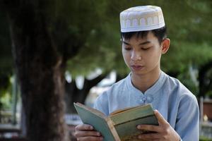 giovane asiatico musulmano ragazzo seduta nel scuola parco e lettura il suo libro nel il suo gratuito volte prima andando indietro casa, morbido e selettivo messa a fuoco. foto