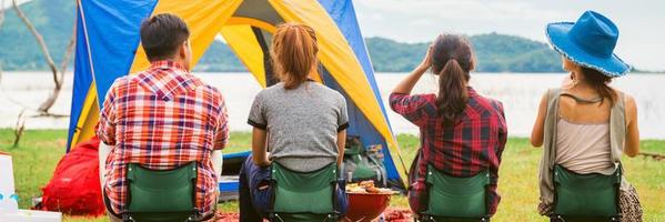 gruppo di uomo e donna godere campeggio picnic e barbecue a lago con tende nel sfondo. giovane misto gara asiatico donna e uomo. panoramico striscione. foto