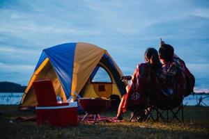 tenda spensierato coppia rilassante a distanza viaggio concetto - romantico asiatico coppia turisti seduta a un' fuoco di bivacco vicino tenda, abbracciare ogni altro sotto alberi e notte cielo. notte campeggio foto