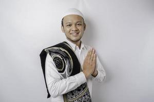 sorridente giovane asiatico musulmano uomo con preghiera tappeto su il suo spalla, Esprimere a gesti tradizionale saluto isolato al di sopra di bianca sfondo foto