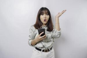 scioccato asiatico donna indossare saggio verde camicia puntamento a il copia spazio su superiore di sua mentre Tenere sua Telefono, isolato di bianca sfondo foto