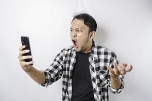 il arrabbiato e pazzo viso di asiatico uomo nel tartan camicia mentre Tenere il suo Telefono su isolato bianca sfondo. foto