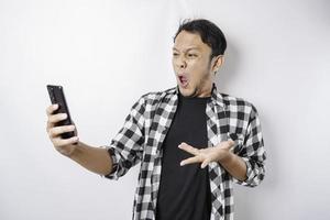 il arrabbiato e pazzo viso di asiatico uomo nel tartan camicia mentre Tenere il suo Telefono su isolato bianca sfondo. foto