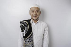 un' ritratto di un' contento asiatico musulmano uomo con preghiera tappeto su il suo spalla è sorridente isolato di bianca sfondo foto