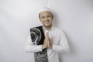 sorridente giovane asiatico musulmano uomo con preghiera tappeto su il suo spalla, Esprimere a gesti tradizionale saluto isolato al di sopra di bianca sfondo foto