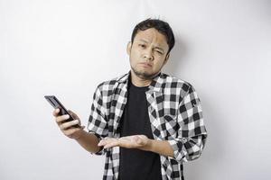 un' insoddisfatto giovane asiatico uomo sembra scontento indossare tartan camicia irritata viso espressioni Tenere il suo Telefono foto