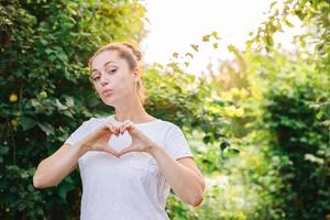 giovane donna sorridente all'aperto. bellissimo bruna ragazza mostrando cuore cartello con mani su parco o giardino verde sfondo. gratuito contento donna a estate. la libertà felicità spensierato contento persone concetto. foto