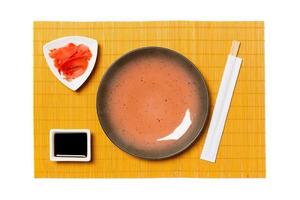 vuoto il giro Marrone piatto con bastoncini per Sushi, Zenzero e soia salsa su giallo bambù stuoia sfondo. superiore Visualizza con copia spazio per voi design foto
