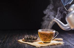 versare un' caldo erbaceo teiera in un' bicchiere tazza, tazza da tè, e asciutto tè le foglie nel un' di legno cucchiaio e posto esso su un' nero di legno tavolo contro un' buio sfondo.