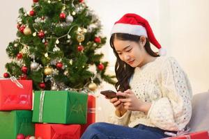 Natale concetto, asiatico donna si siede nel Natale decorato vivente camera e chat su smartphone