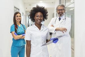 un' multietnico gruppo di tre medici e infermieri in piedi nel un' ospedale corridoio, indossare scrub e cappotti. il squadra di assistenza sanitaria lavoratori siamo fissando a il telecamera e sorridente foto